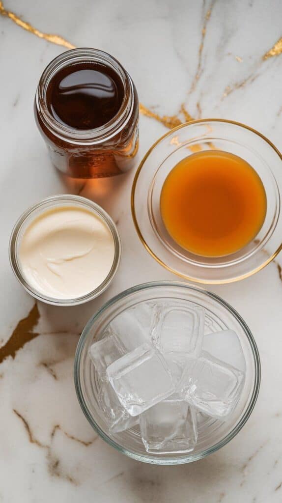 all ingredients for butterscotch bliss dirty soda: a glass bottle of root beer with condensation, thick golden butterscotch syrup in a glass ramekin, heavy cream in a small measuring cup, sweetened condensed milk in a spoon, and ice cubes spilling out of a bowl. Tools include two clear glass cups and a long stirring spoon. 