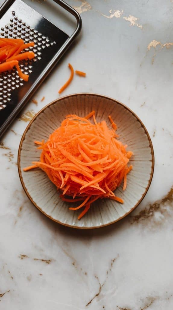 a freshly shredded carrot on a small plate, bright orange strands piled neatly. A grater is placed beside it with a few remaining carrot shreds