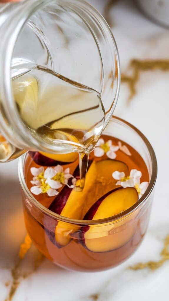 clear elderflower syrup being poured from a glass jar into the plum and gin mixture, syrup glistening as it hits the liquid
