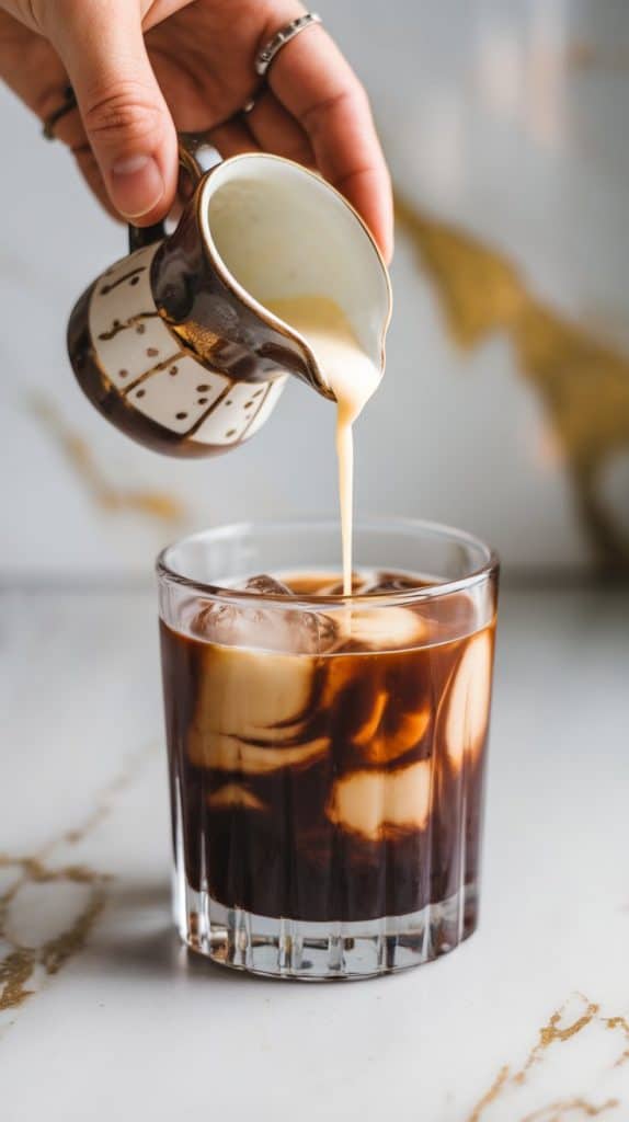 A rocks glass filled with ice as vodka, coffee liqueur, and fresh espresso are poured in, with a small pitcher of cream and coffee beans nearby on a white marble counter.